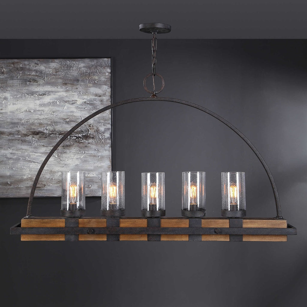 Room setting view Atwood Hanging Island 5 Light Heavy gauge real wood linear body, finished in a mid-tone, featuring clear seeded glass shades. A nod to industrial design is found in the iron wrapped frame, and top arch. The metal work is in a deep weathered bronze finish with a slight texture and black antiquing.
