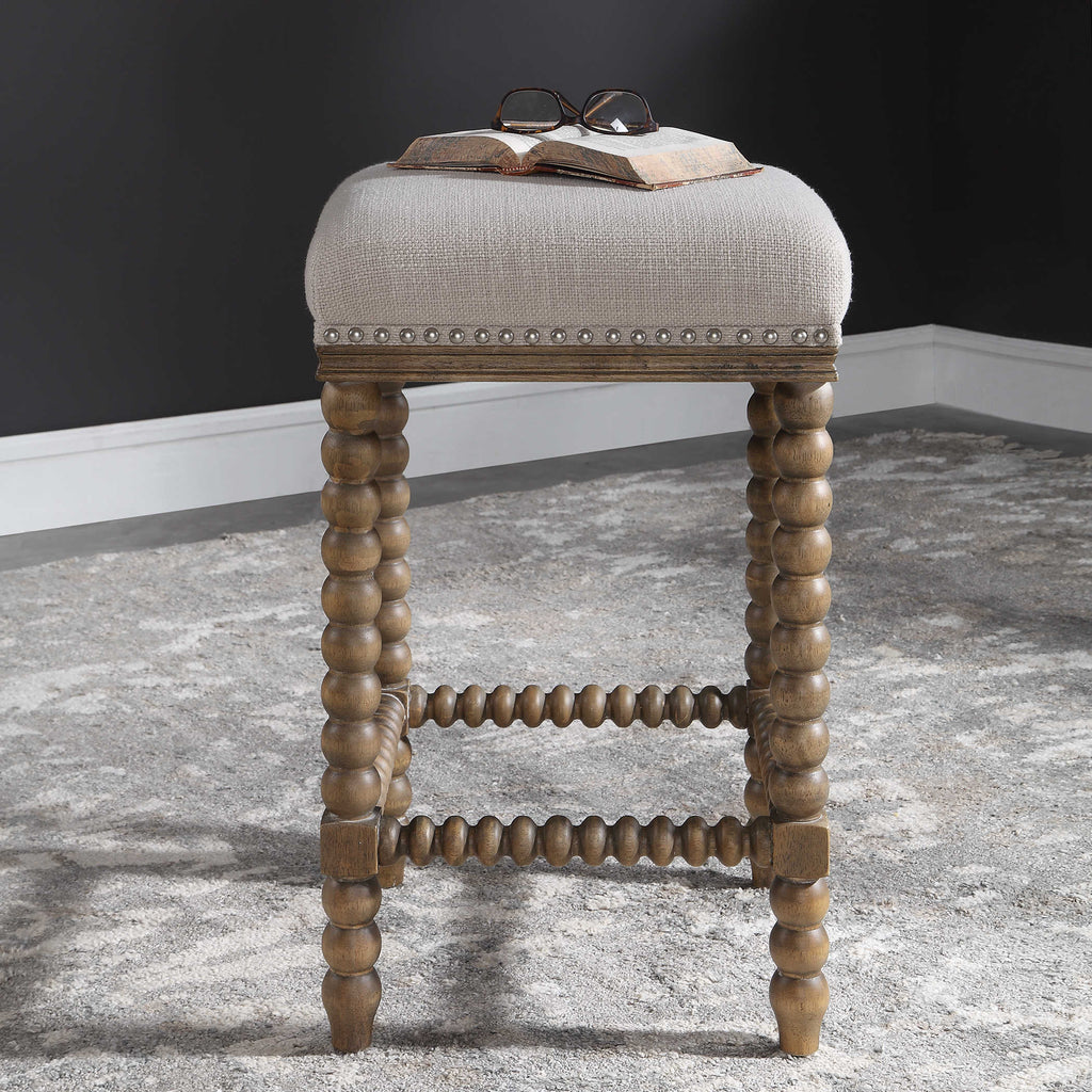 Pryce Counter Stool by Uttermost featuring spindle turned legs and carved trim finished in a light walnut stain. Plush seat is tailored in a soft ivory linen blend fabric accented with brushed nickel nail head trim. Seat height is 26". Room setting view.